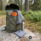 Olivgrön ryggsäck på en klippa i tallskogen med ett grått myggnät som hänger ut över kanten och en orange förvaringspåse som sticker upp ur ryggsäcken, bredvid en vattenflaska i metall och en grå halsduk