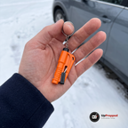 Hand som håller en orange bältesskärare med glaskross och nyckelring framför en snöig gårdsplan