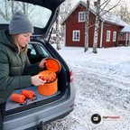 Kvinna i grön dunjacka lyfter locket på den orange väskan med varningsljus i ett öppet bagageutrymme på en grå kombi, två orange nödsovsäckar ligger på filtmattan på en snöig gårdsplan vid en röd stuga