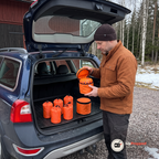 Man i rostbrun arbetsjacka lyfter locket på en orange väska med varningsljus i ett öppet bagageutrymme på en mörkblå kombi, fem orange nödsovsäckar står på gummimattan på en grusgård vid en röd lada