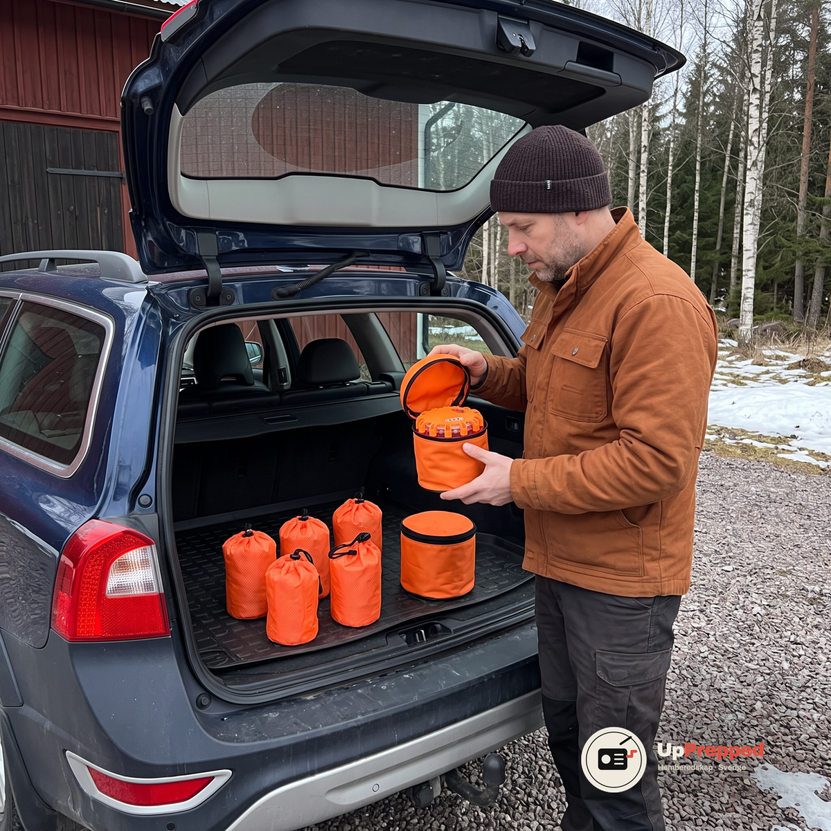 Man i rostbrun arbetsjacka lyfter locket på en orange väska med varningsljus i ett öppet bagageutrymme på en mörkblå kombi, fem orange nödsovsäckar står på gummimattan på en grusgård vid en röd lada