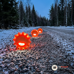 Tre orange LED varningsljus i rad längs en tom grusväg i skymningen, lysande med röd-orange och vita lysdioder bland snö, gran och björk