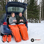 Kvinna och man sitter på bagagekanten av en mörkblå kombi på en snötäckt rastplats med varsin orange nödsovsäck över benen och en mugg i händerna, två orange förvaringspåsar ligger på snön framför dem