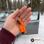 Hand som håller en orange bältesskärare med glaskross och nyckelring framför en snöig parkering vid skogen