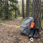 Grå dagsryggsäck mot en tallstam med den orangea förvaringspåsen i sidofickan och ett par vandringskängor bredvid