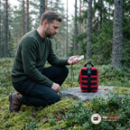Man knäböjer på mossa i en svensk skog med den röda väskan på en granitklippa bredvid sig och en kompass i handen