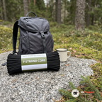 Svart hank paracord på en sten bredvid en grå ryggsäck och en emaljmugg vid en skogsstig