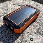 Powerbanken ligger på en solvarm granitklippa med solcellen, den orange ramen och de två runda LED-lamporna i ena änden