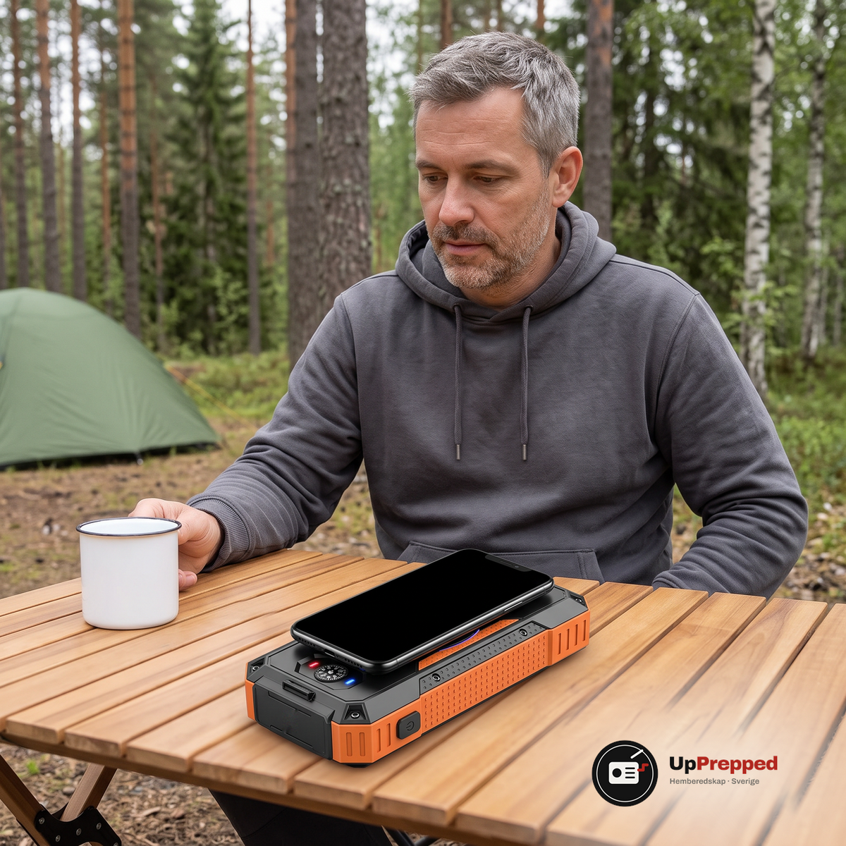 Man i grå munkjacka sitter vid ett trä campingbord i skogen med en svart mobil liggande på laddytan på powerbankens baksida och en vit emaljmugg bredvid