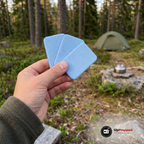 Hand som håller tre ljusblå refillmattor framför ett grönt tält och en kokare på en rastplats i skogen