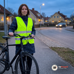 Kvinna i mörkblå regnjacka med reflexvästen över står med sin svarta cykel på en cykelväg vid en villagata i skymningen