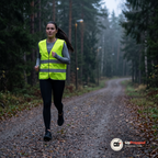 Kvinna med hästsvans springer på en grusväg i skogen i skymningen med reflexvästen över en grå tröja