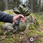 Hand som håller ett grönt regnskydd hopknycklat i näven framför en mossig sten i skogen