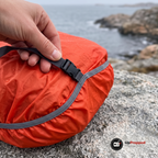 Hand som håller i remmen med spänne på ett orange regnskydd över en ryggsäck på en granitklippa vid havet i lätt regn