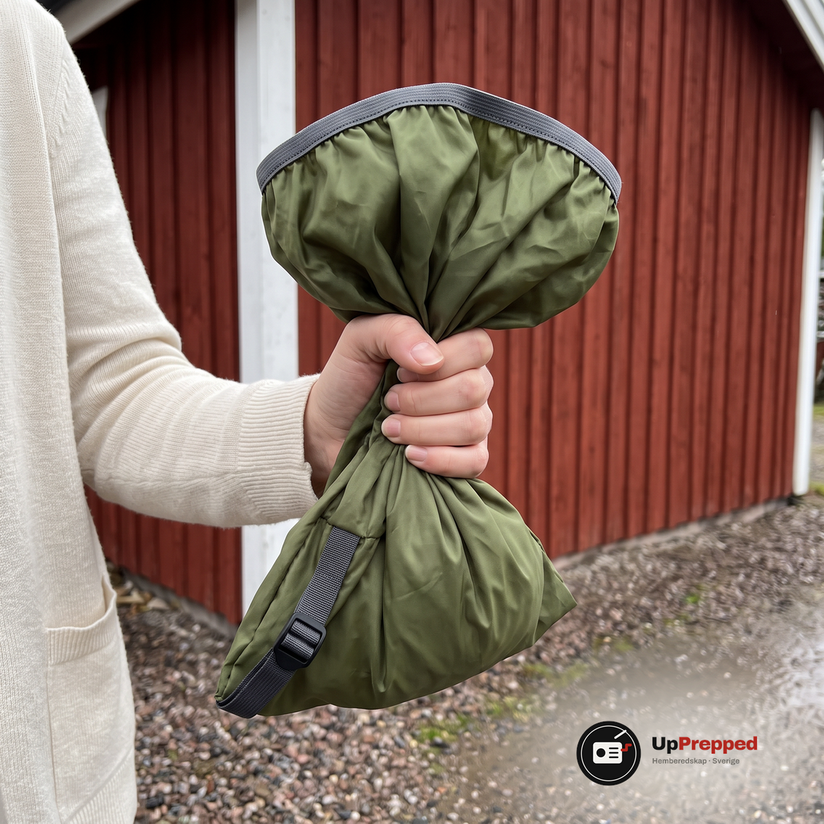 Hand som håller ett grönt regnskydd hopknycklat i nävan framför en rödmålad träskjuls vägg