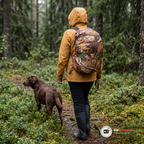 Kvinna i senapsgul regnjacka går med en brun labrador på en blöt skogsstig med en dagsryggsäck som har ett regnskydd i lövkamouflage