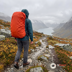 Man i mörkgrön regnjacka går på en stenig fjällstig i regn med en stor ryggsäck som har ett orange regnskydd, sedd bakifrån