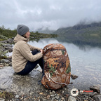 Kvinna i beige jacka sitter på ett granitblock vid en fjällsjö med en ryggsäck som har ett kamouflageskydd bredvid sig