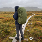 Kvinna i mörkblå regnjacka står på en spång över en myr i svenska fjällen i regn med en stor ryggsäck som har ett grönt regnskydd