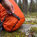 Hand som håller i remmen med spänne på ett orange regnskydd över en stor ryggsäck på en mossig granitsten i en tallskog i lätt regn