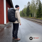 Man med grått skägg står på en regnvåt perrong vid en röd trästation med en stor ryggsäck som har ett svart regnskydd