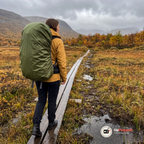 Kvinna i gul regnjacka går på en spång över en myr i fjällen i regn med en stor ryggsäck som har ett grönt regnskydd, sedd bakifrån