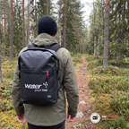 Man i olivgrön skaljacka går på en stig i barrskog med den svarta vattentäta ryggsäcken på ryggen