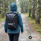 Kvinna i blå regnjacka går längs en grusväg i ösregn med den svarta ryggsäcken på ryggen