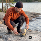 Man i rostorange tröja värmer vatten på spritbrännaren med en liten kastrull på en klippa vid en sjö