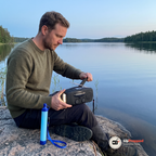 Man vevar nödradion på en sten vid en sjö i kvällsljus med vattenfiltret bredvid sig