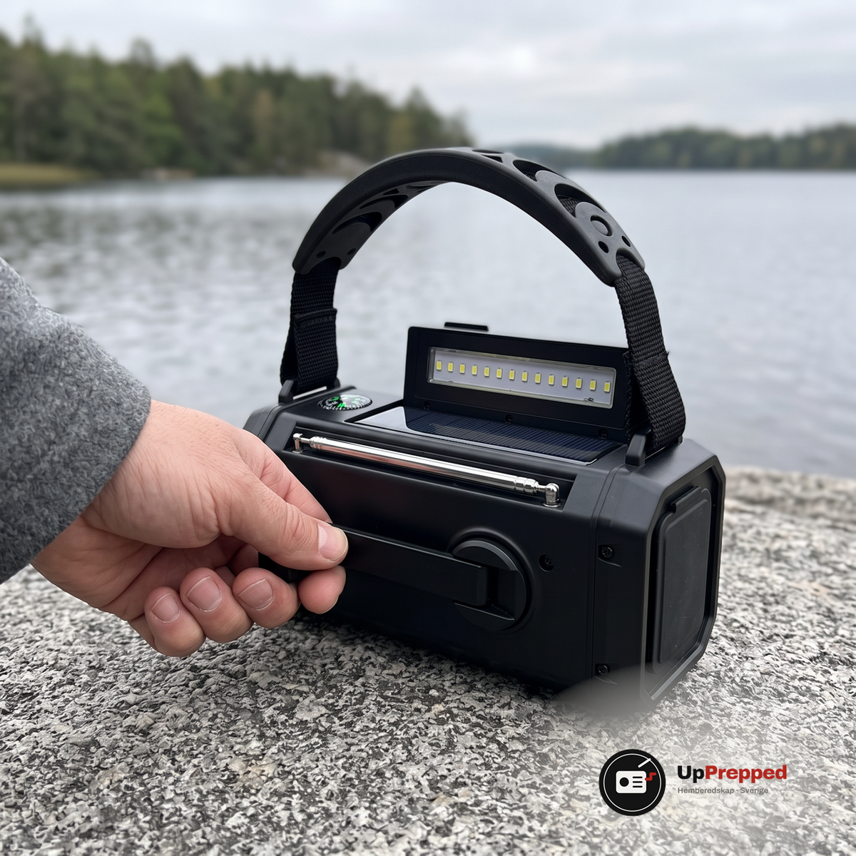 Hand som tar tag i den hopfällda veven på baksidan av nödradion på en sten vid en sjö, med handtag, lampa och antenn