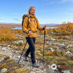 Kvinna i senapsgul skaljacka går på en stenig stig över kalfjället på hösten med en vandringsstav i varje hand