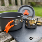 Närbild på kastrullens orangea hopfällbara handtag med silverfärgat beslag och nitar, med locket och spritköket med sitt lock bredvid på ett skifferunderlag