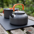 Närbild på vattenkokarens orangea handtag, pip, lock med trådögla och silverfärgat beslag på ett skifferunderlag, med kastrullen suddig bakom
