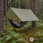 Man i grå ulltröja ligger i en hängmatta under en olivgrön tarp som är uppspänd mellan tallar i skogen