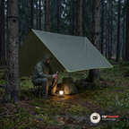 Person i regnjacka sitter på en campingpall under en olivgrön tarp i en regnig granskog med en tänd LED-lykta bredvid