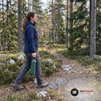 Kvinna går på en stig i tallskog och bär en grön termosflaska i bäröglan