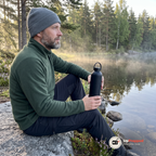Man sitter på en sten vid en svensk skogssjö i morgondimma och håller en svart termosflaska på knät