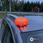 Orange magnetiskt LED varningsljus på taket av en grå kombi i skymningen med röd lada och skog bakom