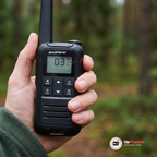 Närbild på en walkie talkies display och knappar i en hand framför en suddig skog