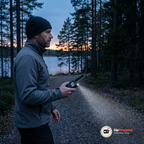Man i grå jacka och mössa håller en walkie talkie med tänd ficklampa på en grusväg vid en sjö i skymningen