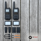 Två walkie talkies, två batteripaket, två bältesclips, två batteriluckor, en laddkabel och två handlovsband på ett grått träbord